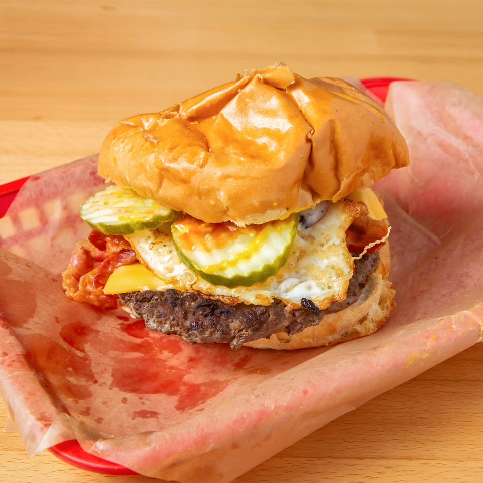 A burger with a fried egg, bacon, cheese, and pickles on a bun in a red basket.